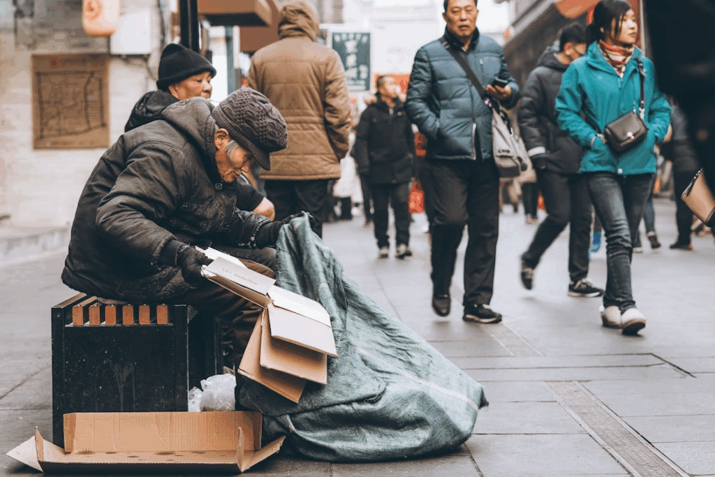 Older adult sitting on a city sidewalk while pedestrians walk past, illustrating aging populations and economic inequality.