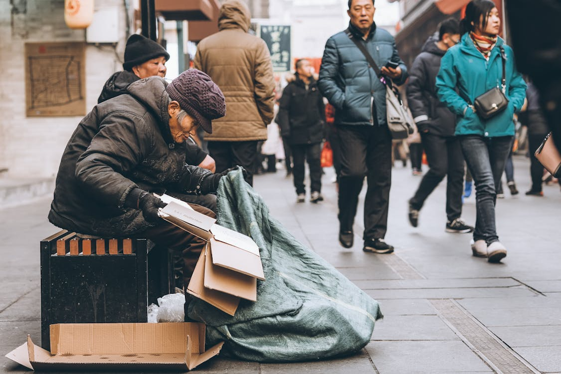 Older adult sitting on a city sidewalk while pedestrians walk past, illustrating aging populations and economic inequality.